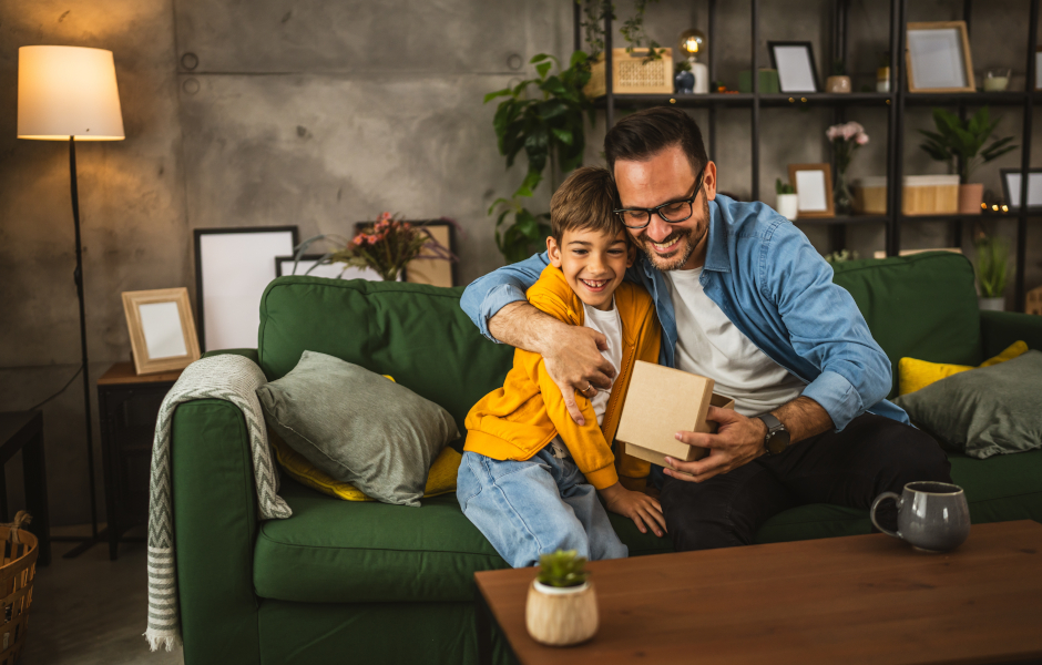 Father and son smiling and hugging on a sofa, father holding a gift for Father's Day
