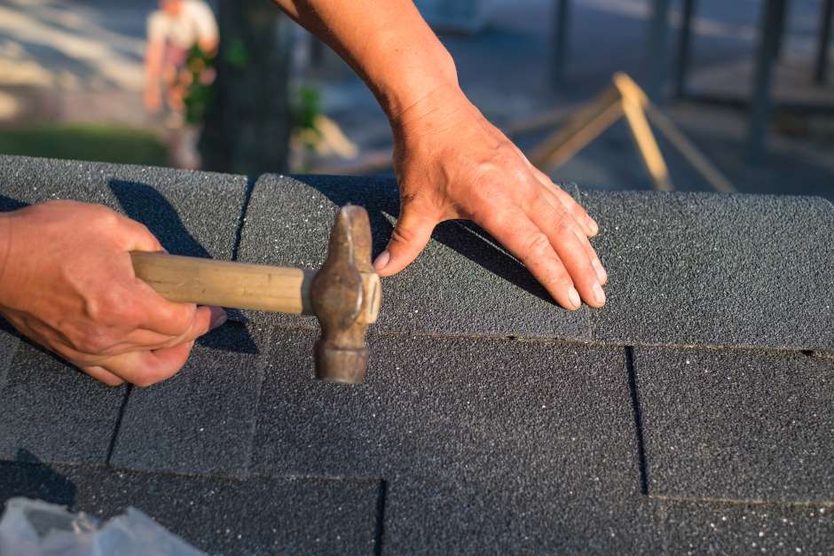 Image of a workers hands, one hand holdind onto a hammer while the other holds a roof shingle